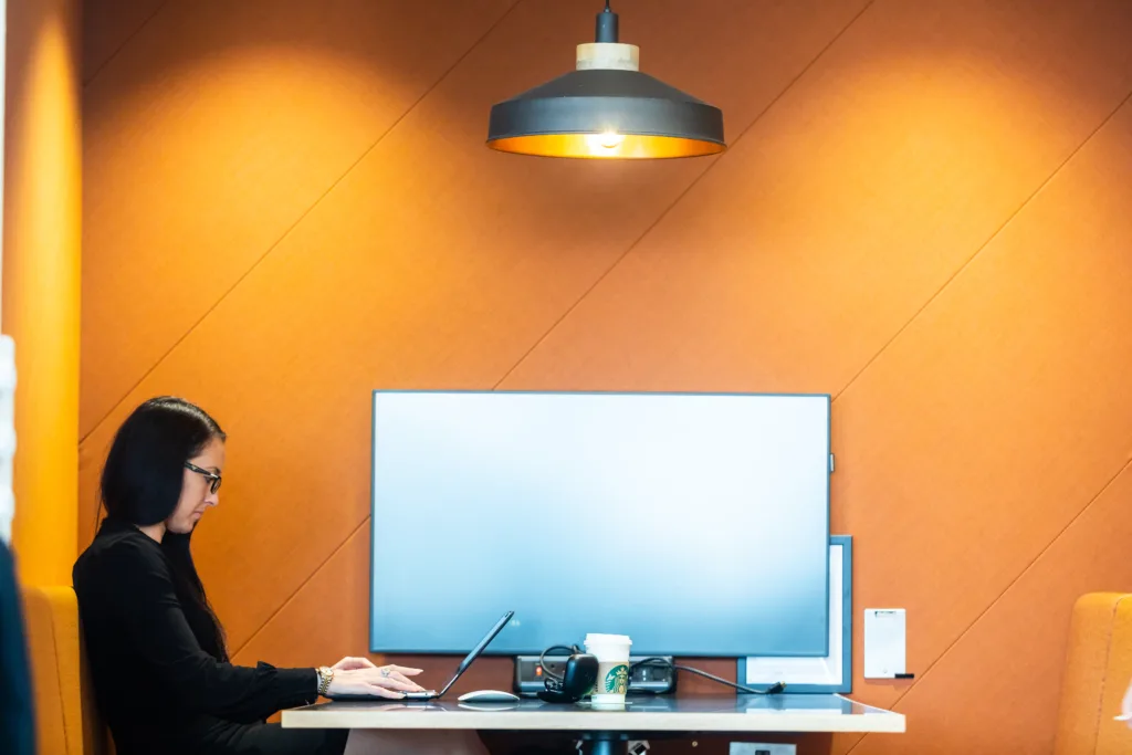Mitie employee working on a laptop in an orange-coloured booth in an office, with a screen on the wall
