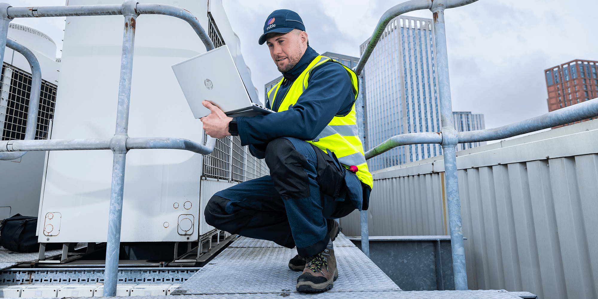Engineer in high-visibility clothing using a laptop beside rooftop mechanical equipment.