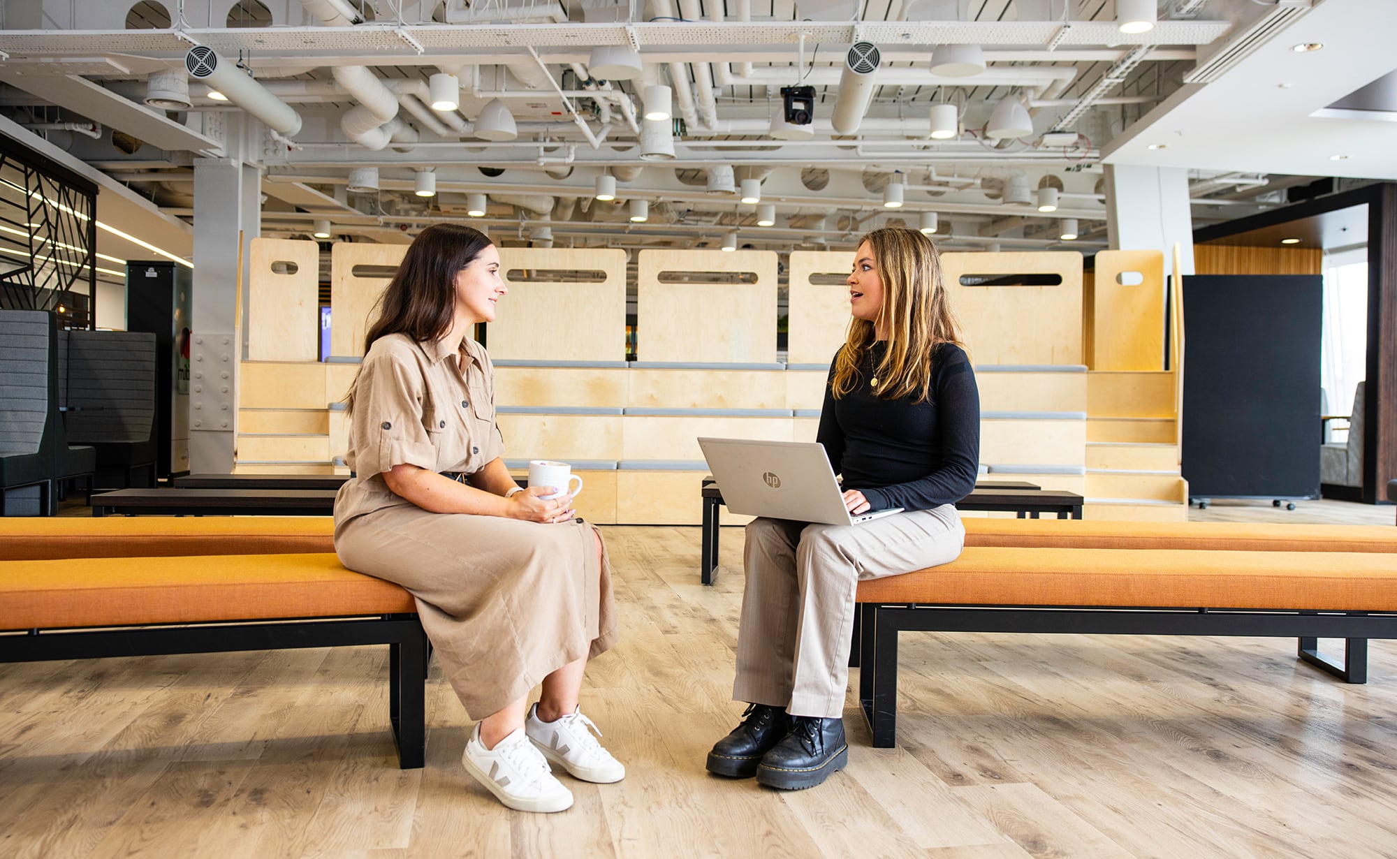 Two colleagues talking in a modern office workspace, one holding a laptop.