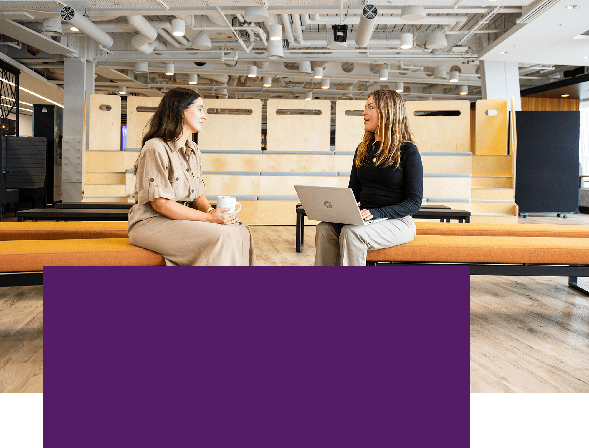 Two colleagues talking in a modern office workspace, one holding a laptop.