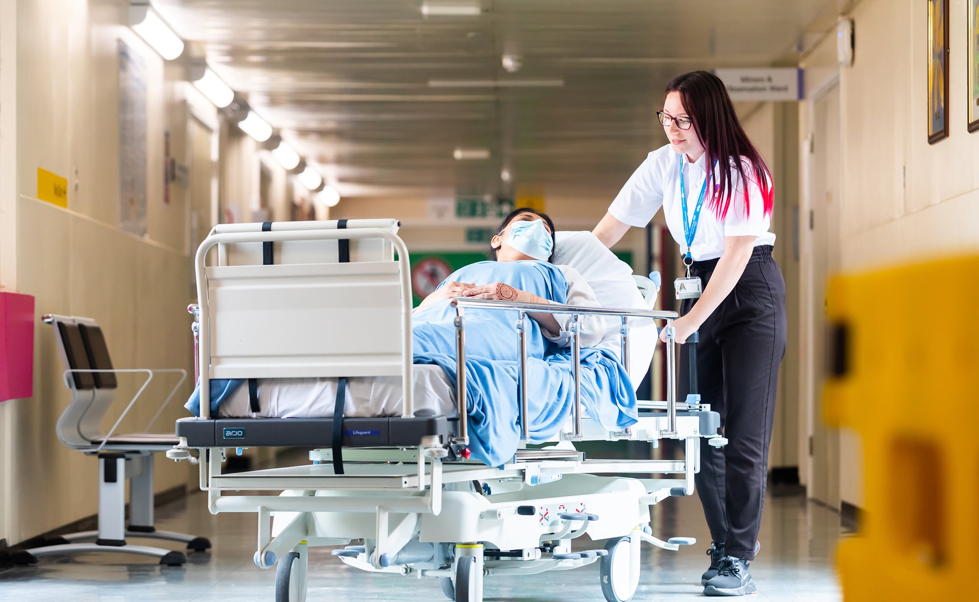 Healthcare worker pushing a patient on a hospital bed along a corridor.